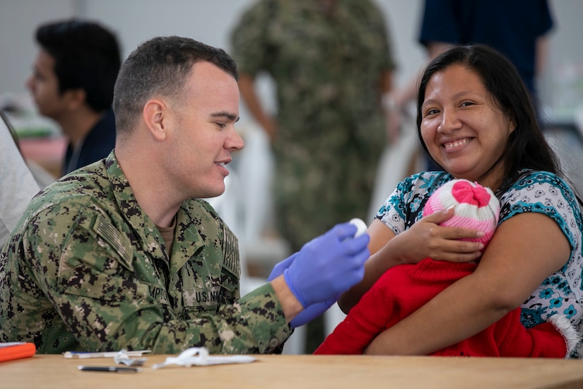 A man wearing a military uniform and surgical gloves treats a baby being held in mother's arms.