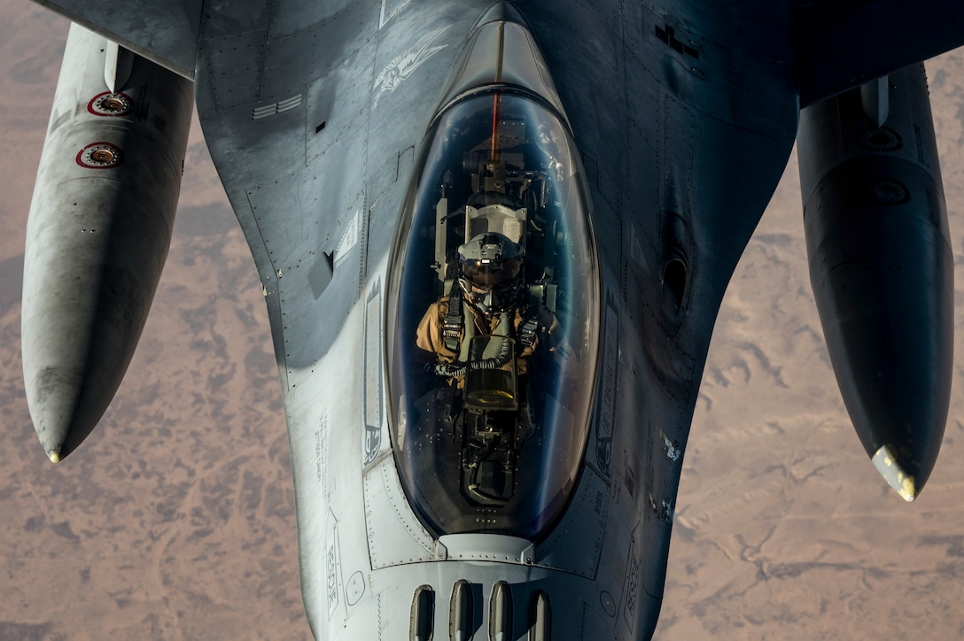 A pilot, shown from overhead, is visible in the cockpit of a jet in flight.