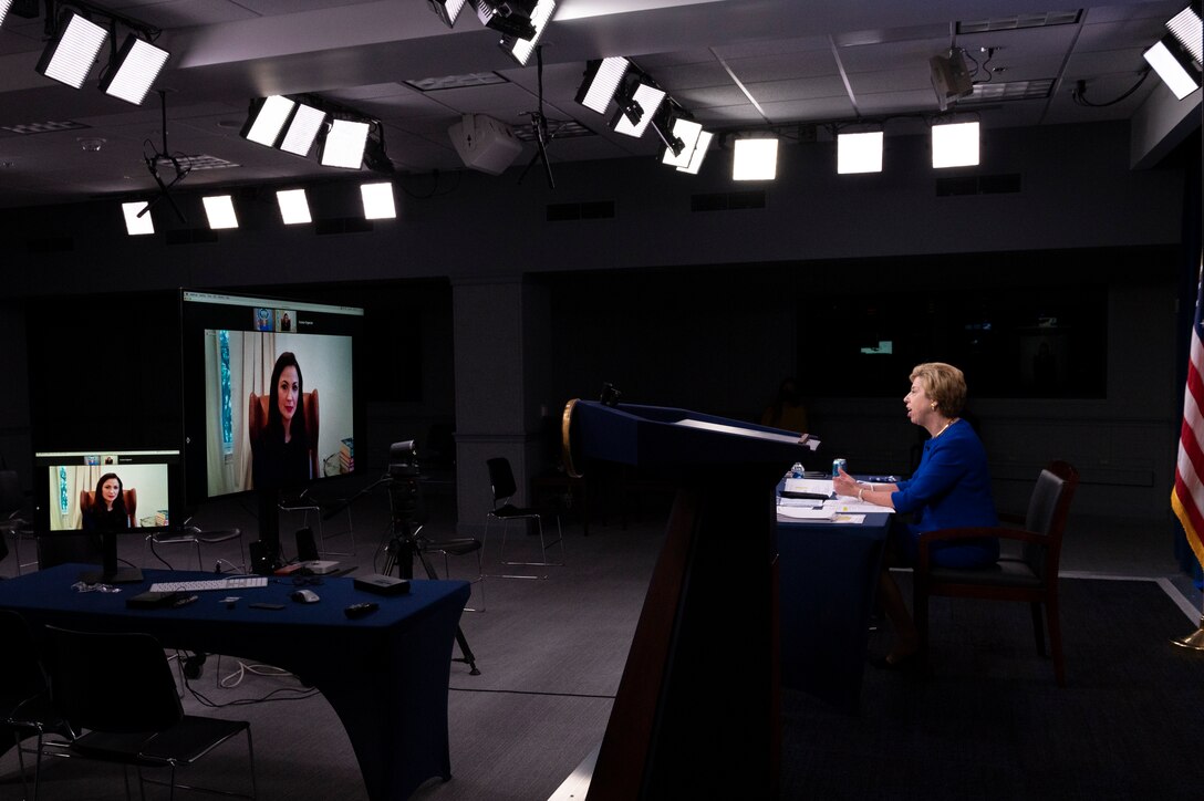 A woman sits at a desk. She looks at a TV screen with the image of another woman on it.