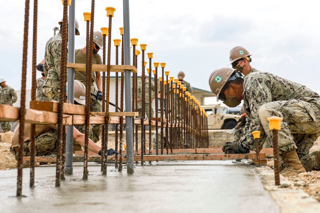 Metal bars stick up from a concrete pad where Navy Seabees wearing hard hats are working.