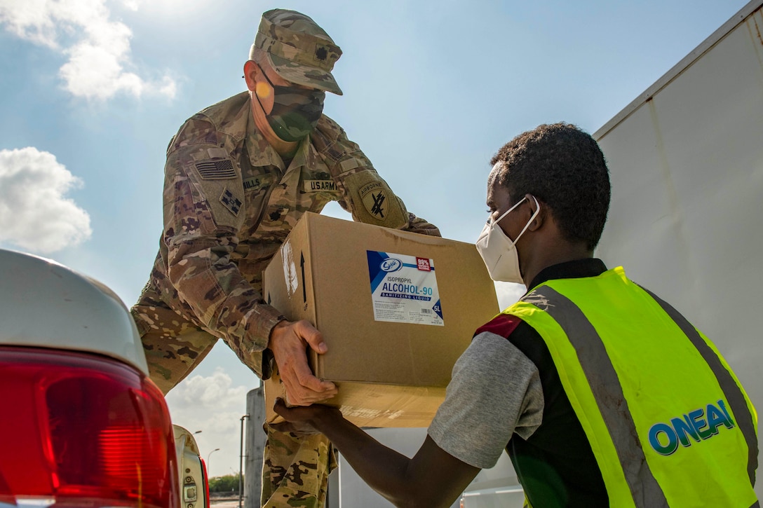 A soldier wearing a face mask passes a box of supplies to a civilian, also wearing a mask.