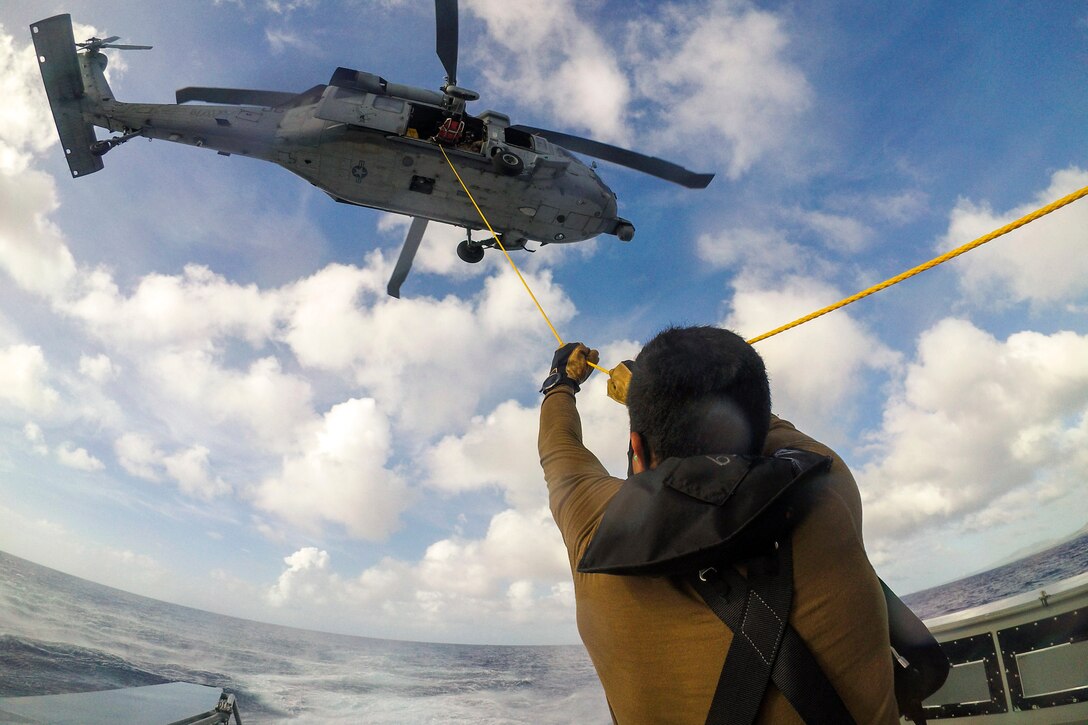 A sailor holds a rope from an airborne helicopter while standing on a ship in a body of water.