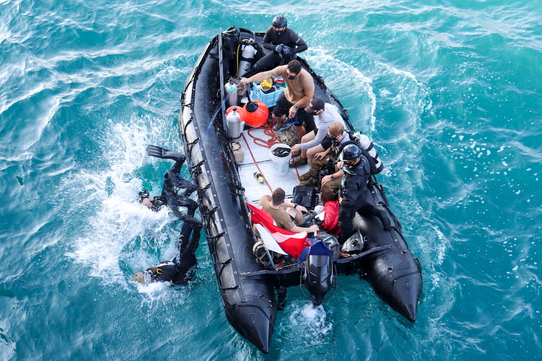 A group of people sit inside a small inflatable boat as two sailors plunge back first into water.
