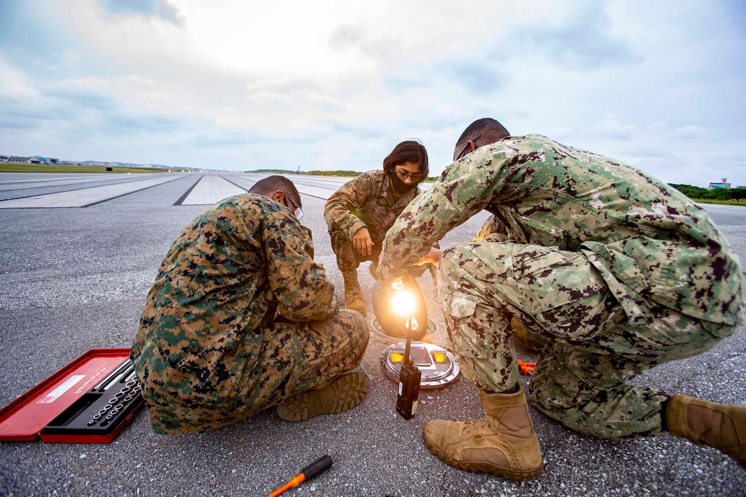 Three Marines repair a light while sitting and kneeling on an airfield.