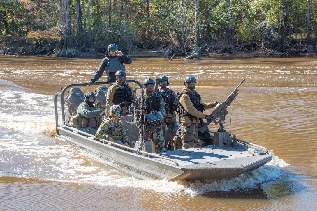 A group of foreign service members ride in a boat through a river; a tree line can be seen behind.