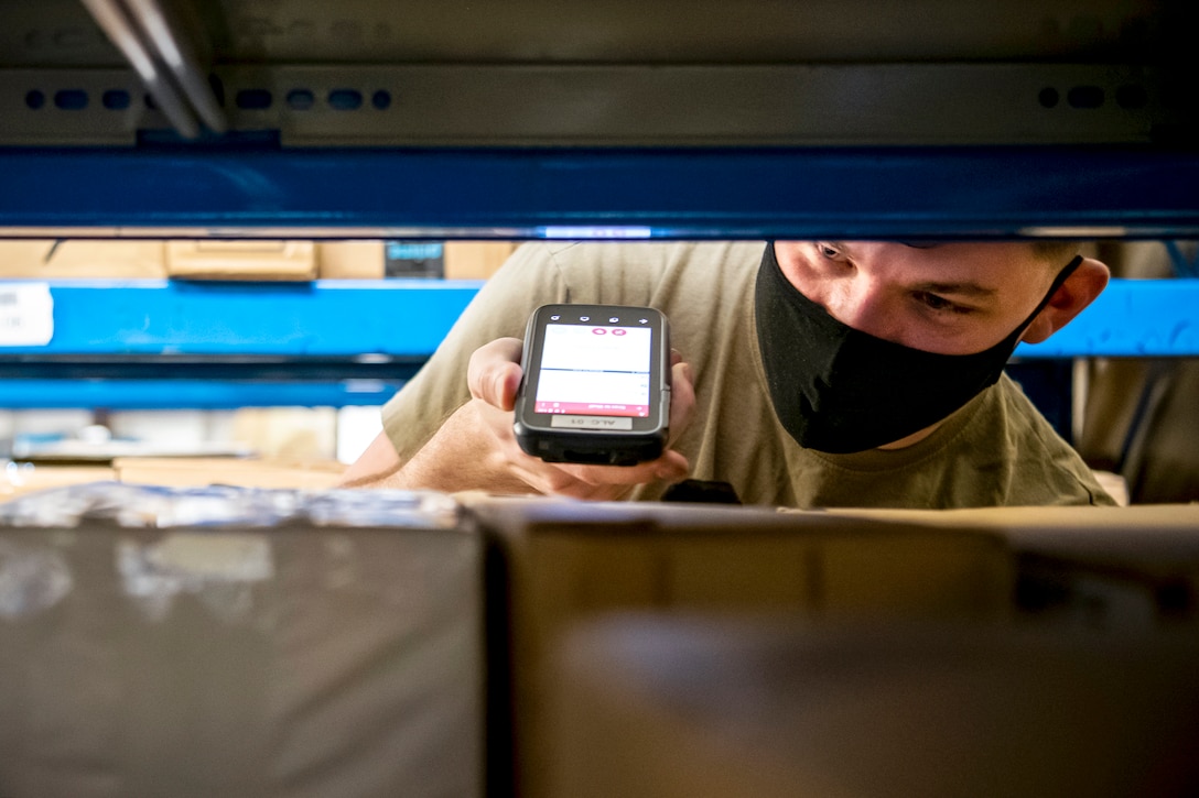 An airman wearing a face mask holds a scanner up to packages on shelves.