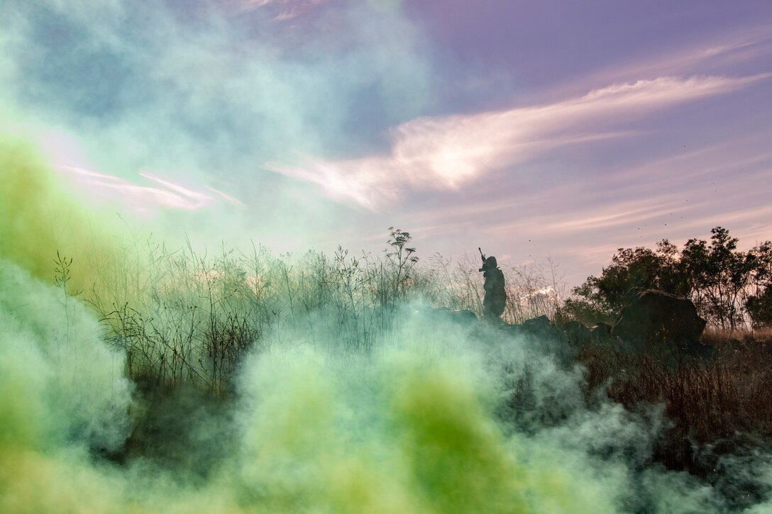 An airman stands in a wooded area surrounded by a cloud of yellow smoke.