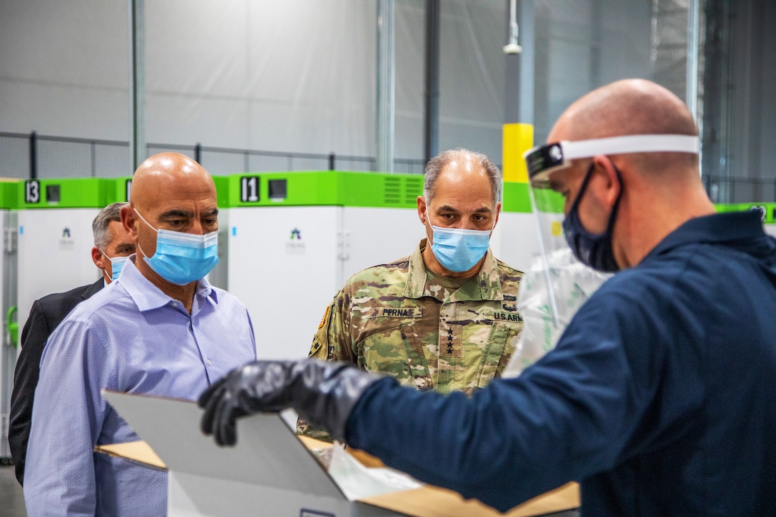 A man in a military uniform and two men wearing civilian clothes watch as another man opens a box. All of the men wear face masks.