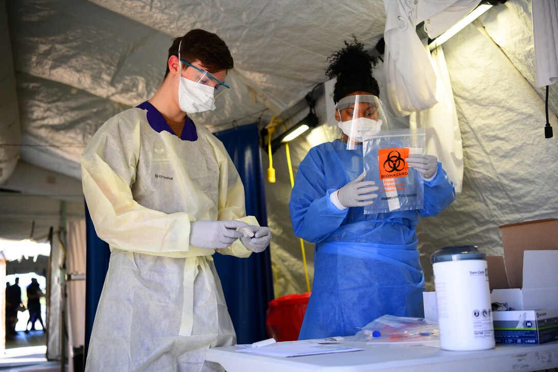 A man and a woman wear personal protective equipment while working inside a tent. The man examines a small tube that he holds in his hand; the woman holds a plastic bag with the word "biohazard" written on the outside.