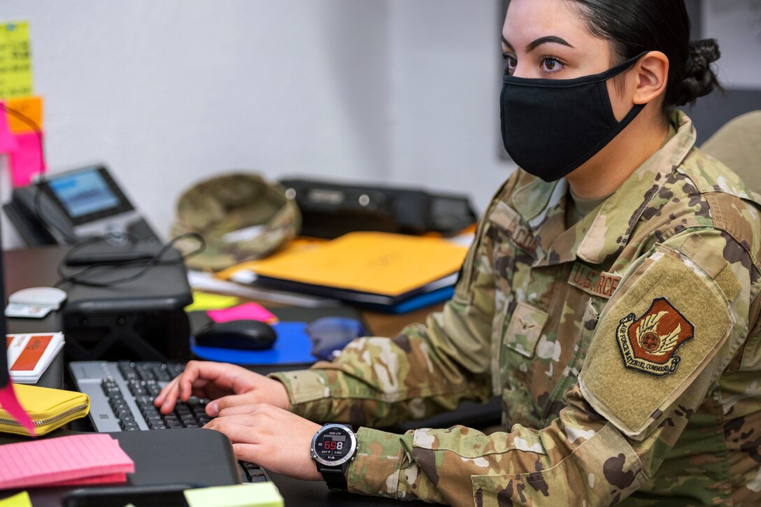 A woman in a military uniform wears a face mask as she types on a keyboard.