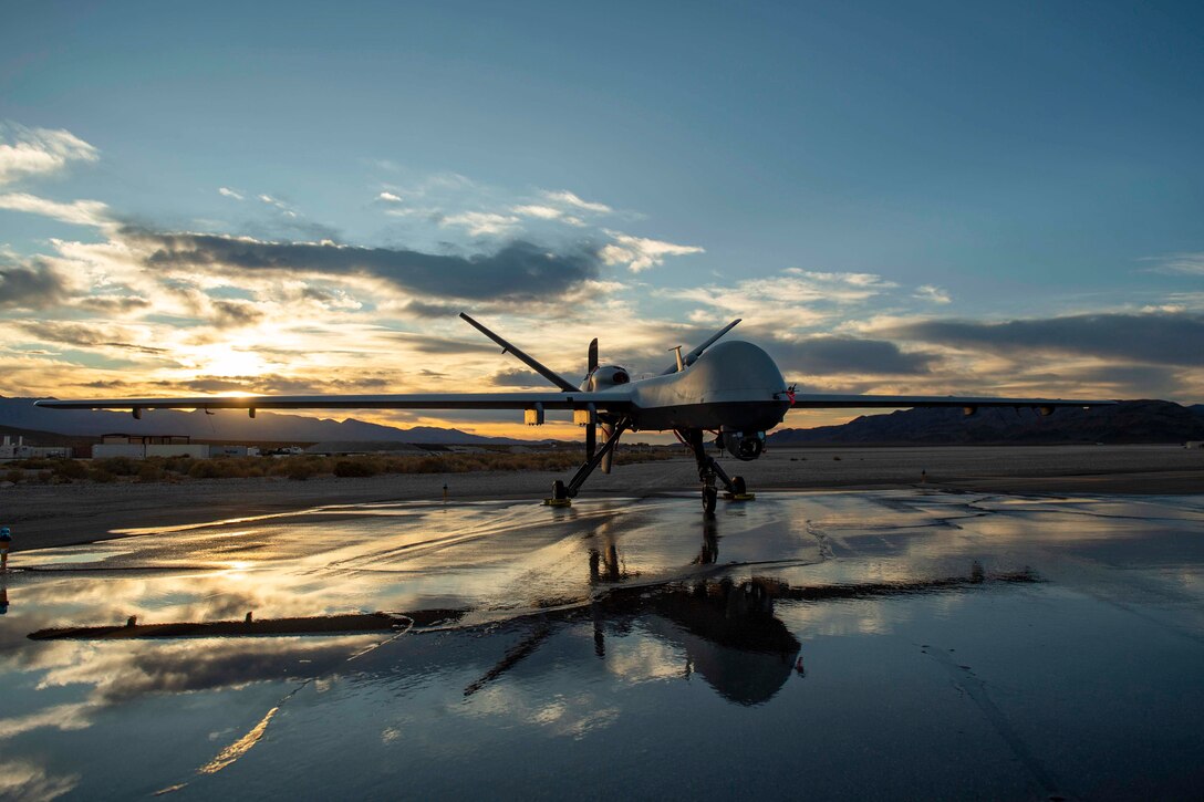 An Air Force MQ-9 Reaper sits parked on a wet parking lot at sunset.