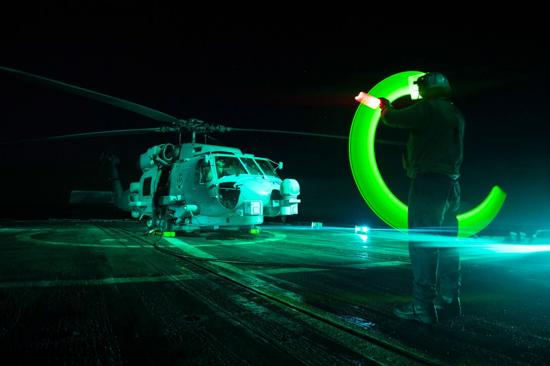 A sailor uses a green light to signal toward a helicopter on the deck of a ship.