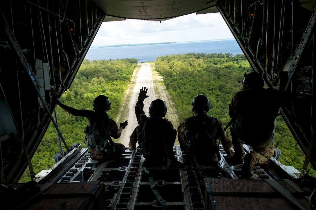 Four airmen, shown from behind in silhouette, wave from the open back of an aircraft flying over an island.