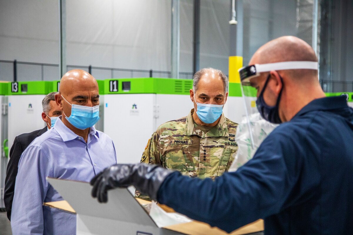 A man in a military uniform and two men wearing civilian clothes watch as another man opens a box. All of the men wear face masks.