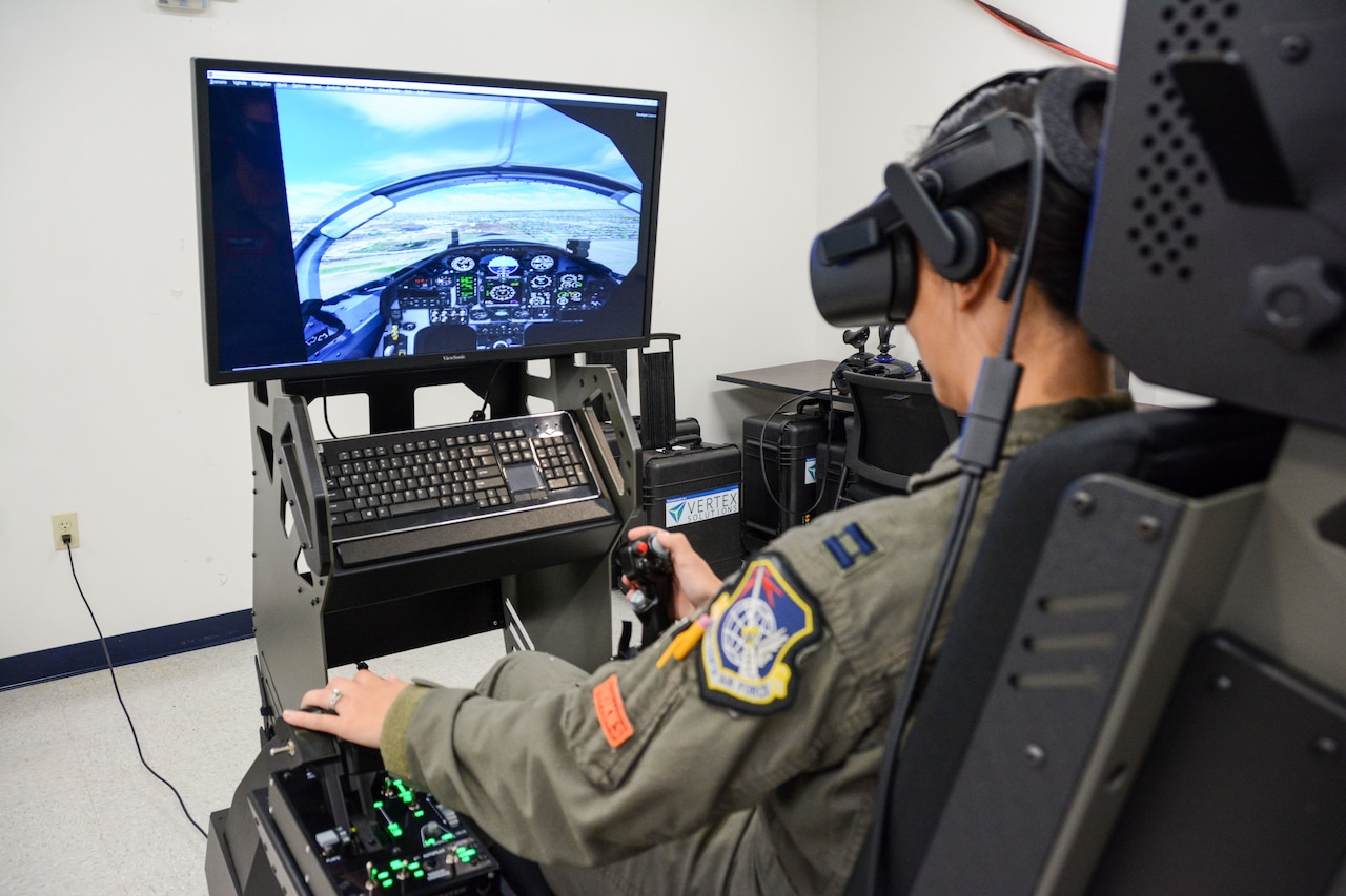 A woman in a military flight suit operates an aircraft simulator. A replica of the  cockpit can be seen on the flight simulator monitor.