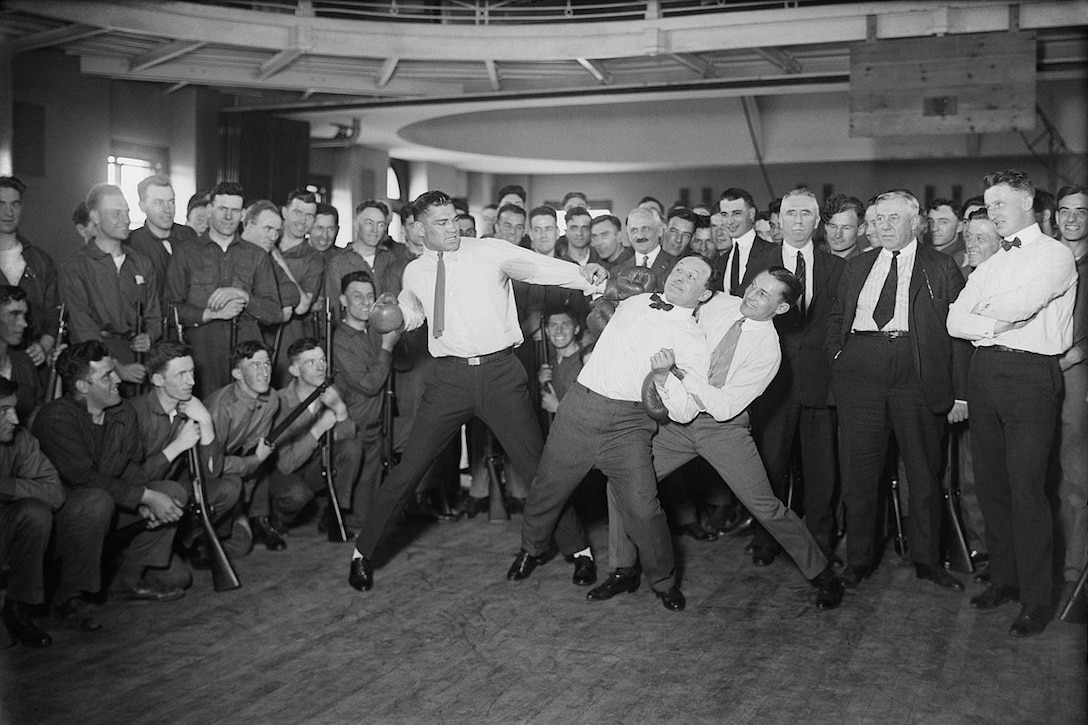 Two men wearing dress shirts, ties and boxing gloves pretend to fight in front of a crowd. A third man, who is also wearing a dress shirt and tie,  holds the arms of one of the men.