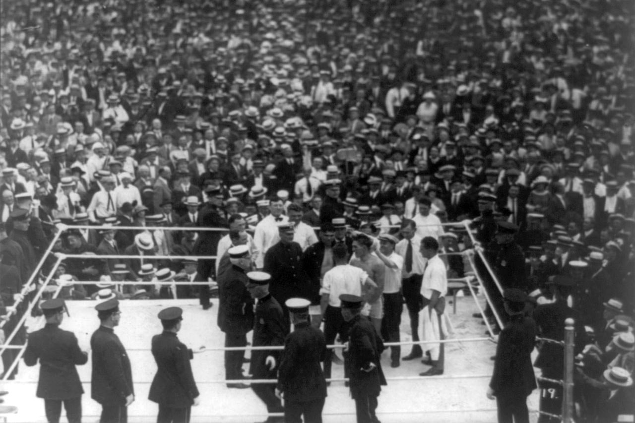 A photo provides an aerial view of a group of people in a boxing ring surrounded by a large crowd.