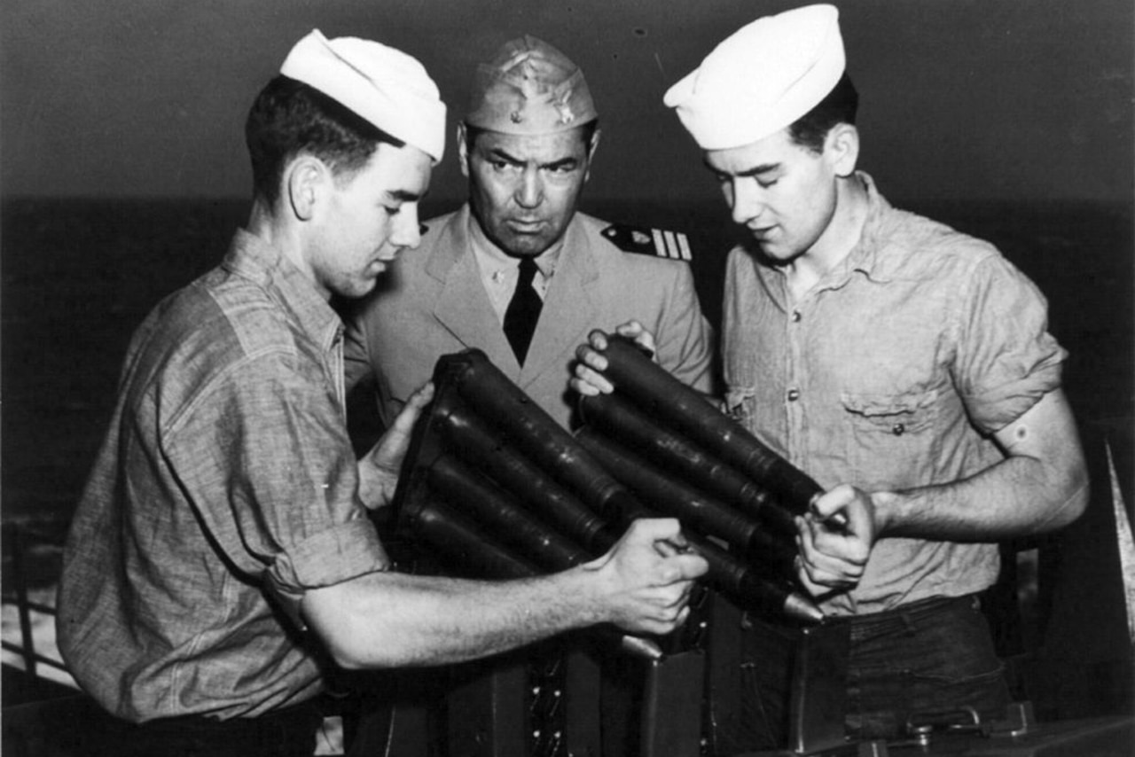 Two young men in sailor uniforms load an antiaircraft gun while an older man in an officer’s uniform looks on.