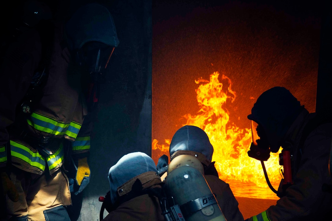 Four sailors wearing fire protection gear stand in a doorway in front of a fire.