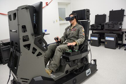 A woman in a military flight suit operates an aircraft simulator. A replica of the  cockpit can be seen on the flight simulator monitor.