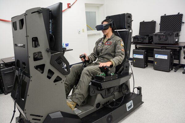 A woman in a military flight suit operates an aircraft simulator. A replica of the  cockpit can be seen on the flight simulator monitor.
