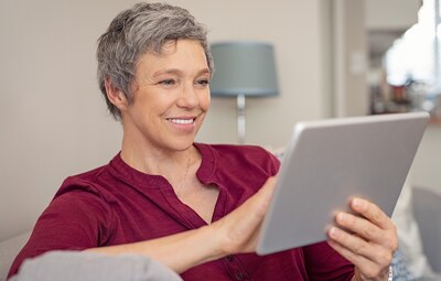 Smiling senior woman looking her digital tablet while sitting on sofa. Portrait of mature happy woman relaxing at home with digital tablet. Happy lady with gray hair browsing on laptop in living room.