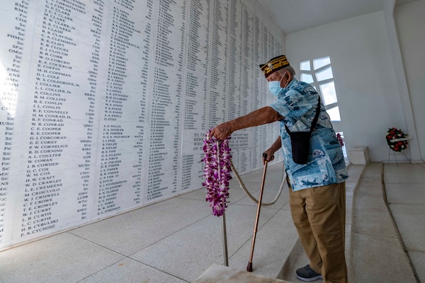 A man places a lei on a metal pole while standing in a room with names carved on the wall.