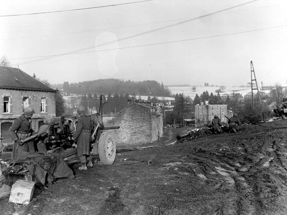 Two men prepare a field artillery gun while two more men crouch down looking in the distance at a town.