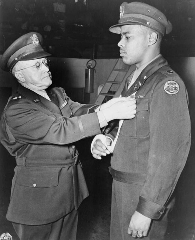 A man in an officer’s uniform pins a medal to the lapel of the uniform worn by a man whose arm is in a sling.