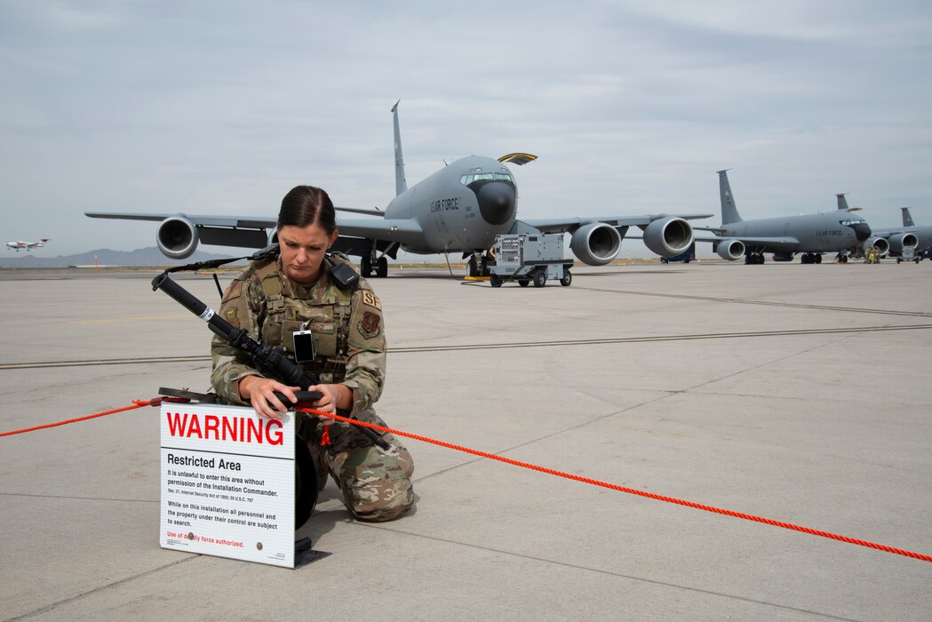 A female Security Forces Airman in Air Force uniform, kneels on a flightline while stretching a red cord out with a KC-135 Stratotanker behind her.
