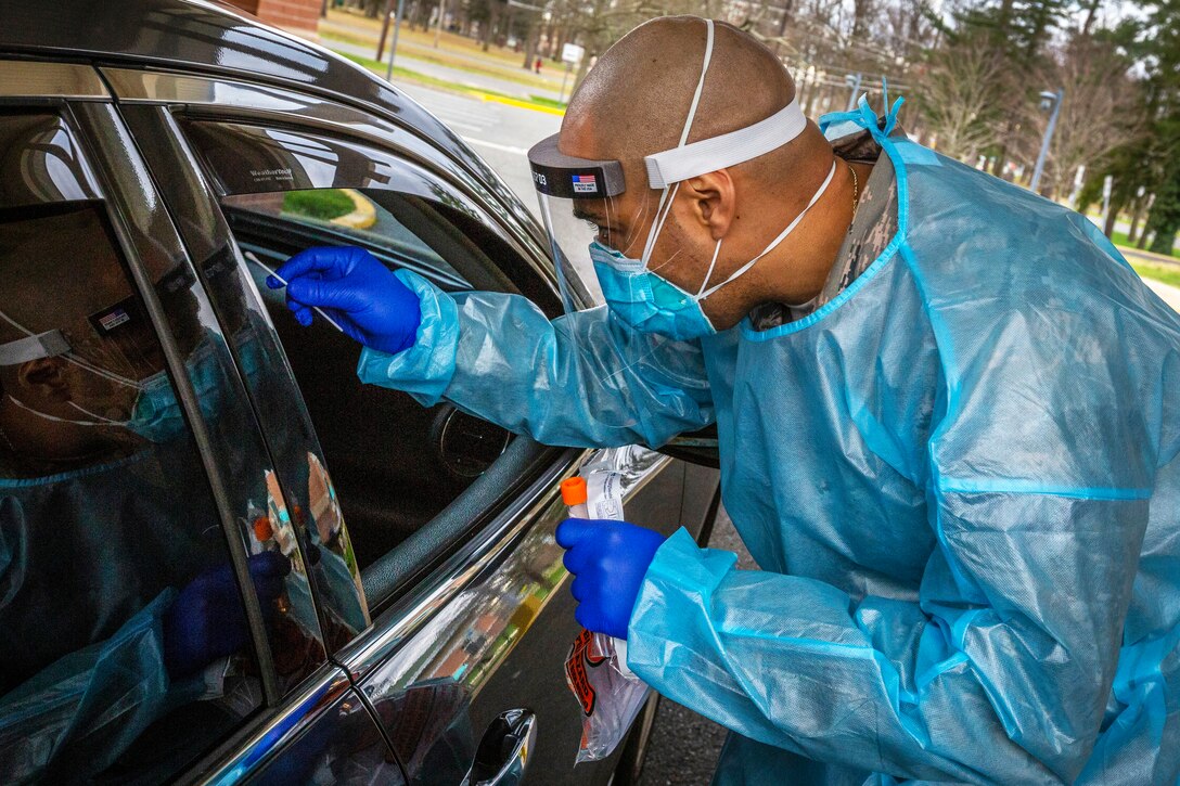 A man wearing personal protective equipment reaches through a car window with a nasal swab.