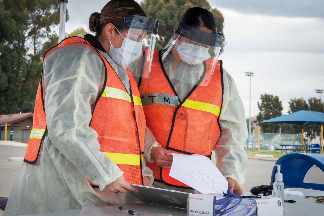 Two women wearing personal protective equipment look at a piece of paper on a table outside.