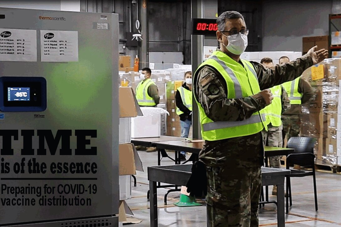 An airman wearing a face mask gestures to his left as he stands inside a warehouse.