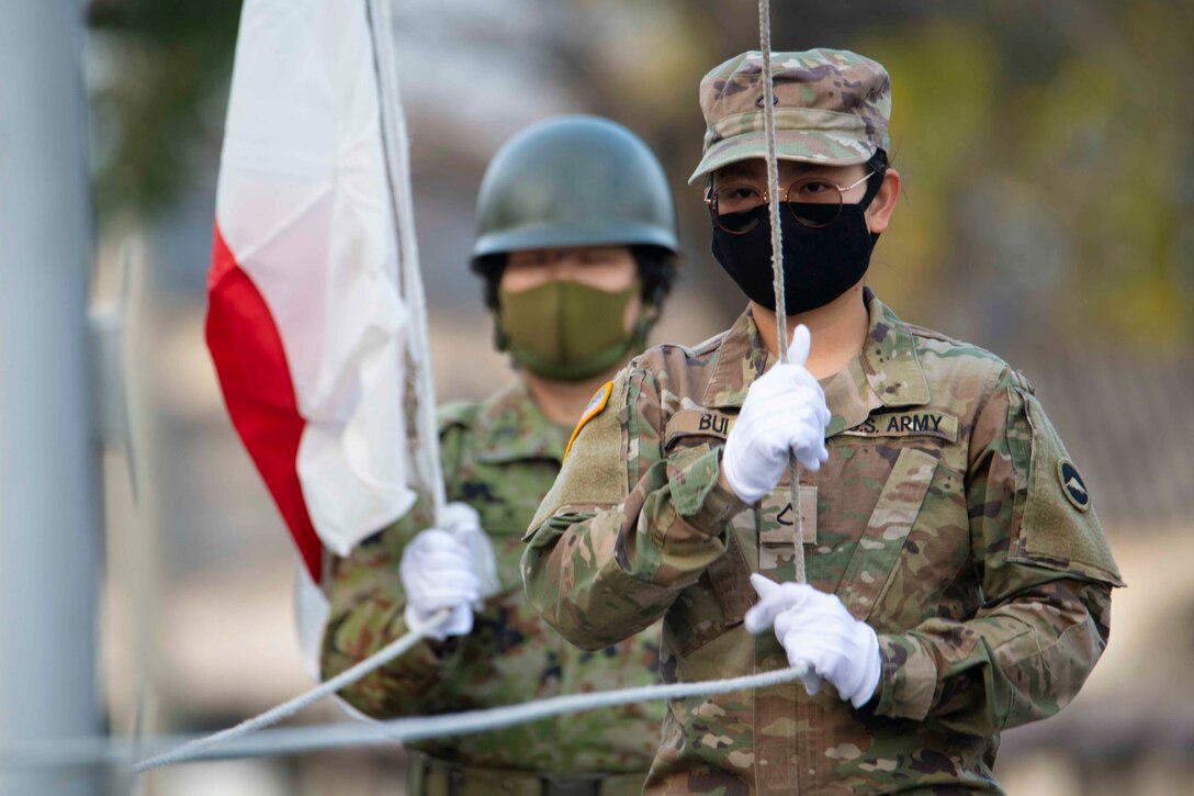 A soldier holds on to a rope while standing in front of a Japanese service member holding a flag.