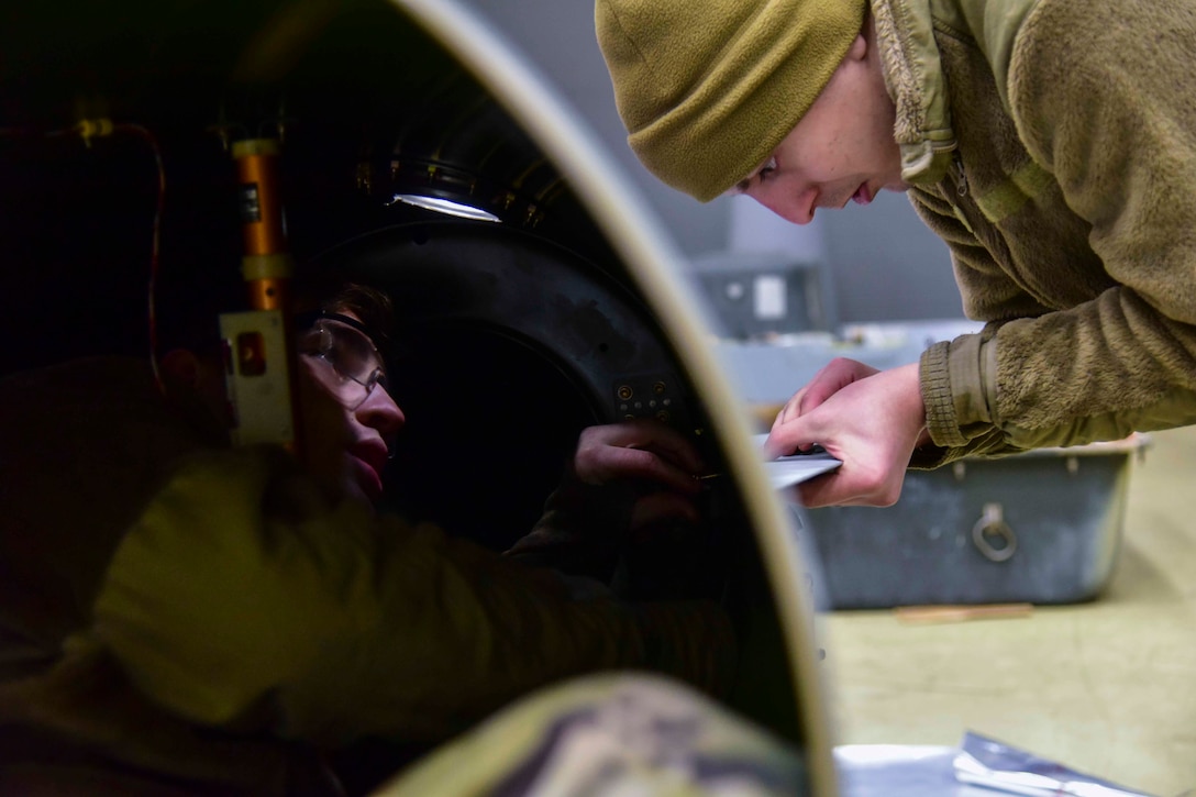 Two airmen work on an external fuel tank; one seen inside the tank and the other stands on the outside.