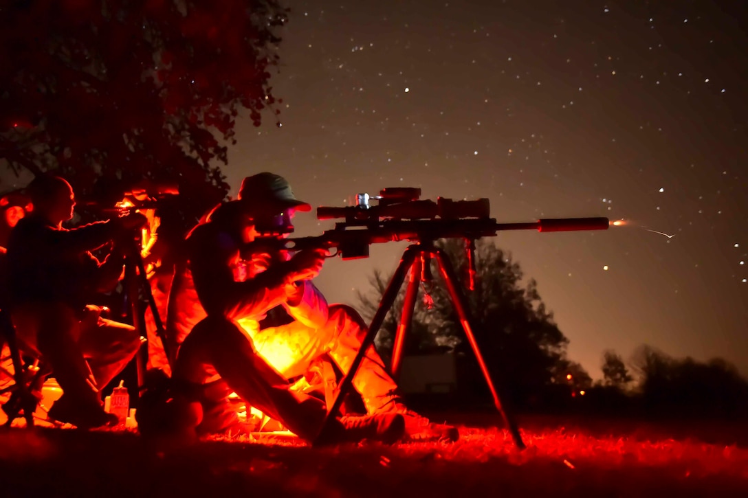 A service member illuminated by red light fires a weapon at night as another person sits behind.