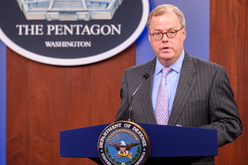 A man standing at a lectern with a microphone speaks. Behind him is a sign indicating that he is at the Pentagon.