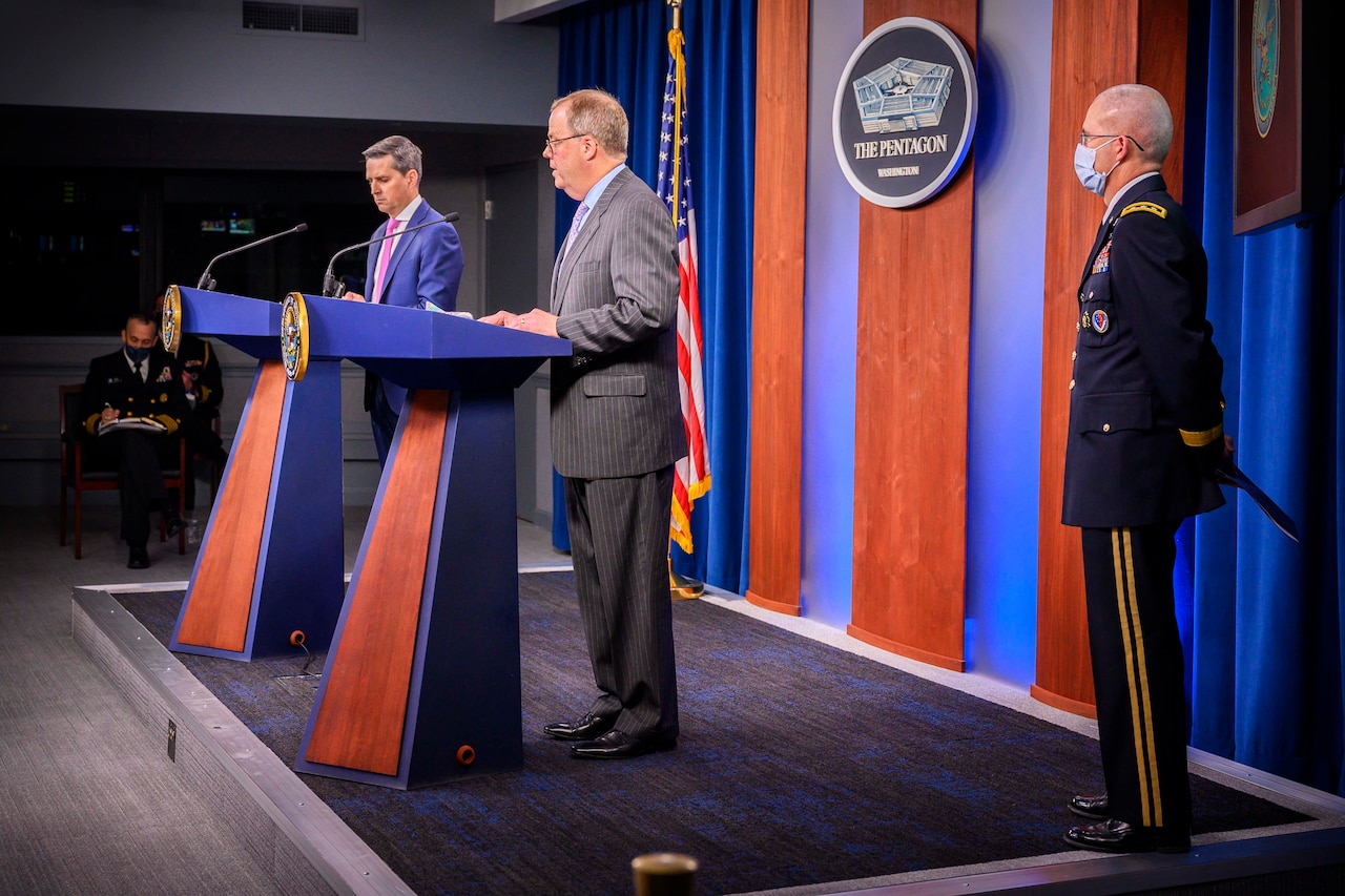 Two men stand at lecterns and speak to a socially distanced audience. A third man in a military uniform stands by.