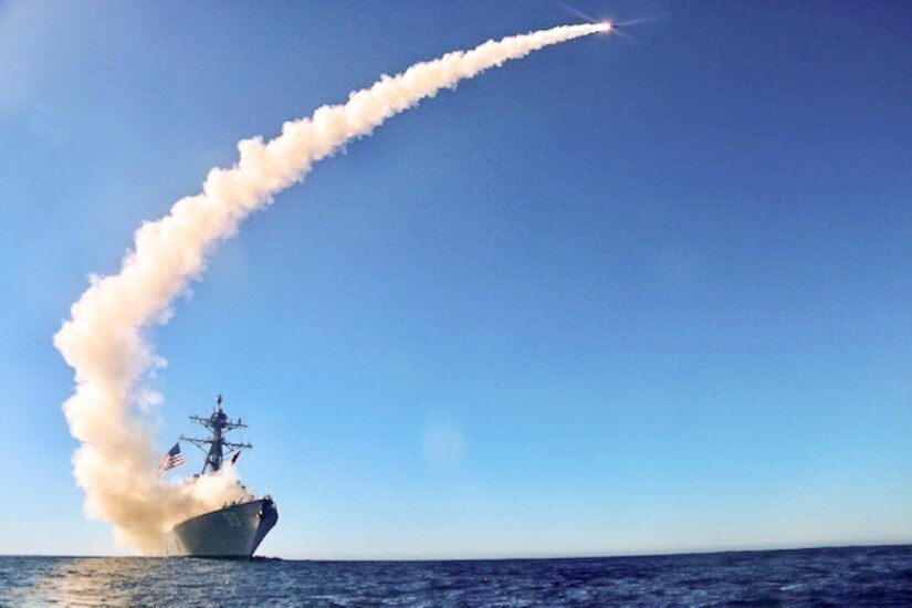 A missile shoots from a ship, created a curved cloud trail in blue sky.