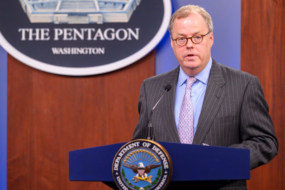 A man standing at a lectern with a microphone speaks. Behind him is a sign indicating that he is at the Pentagon.