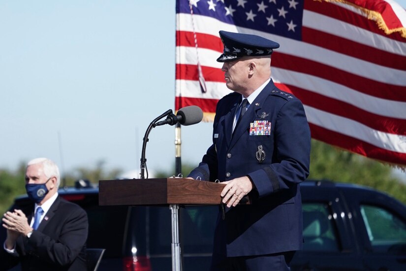 A man in uniform speaks with an American flag flying behind him.