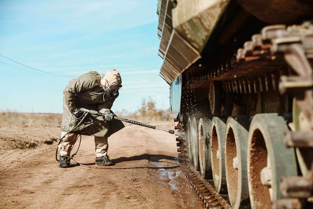 A Marine in personal protective equipment uses a hose to wash the tires of a military vehicle.