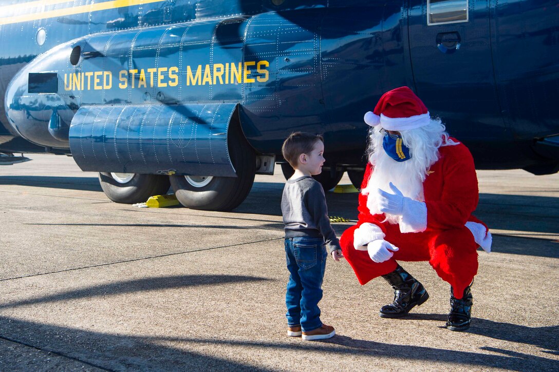 A service member dressed in a Santa costume kneels in front of a child; a Navy aircraft is parked behind.