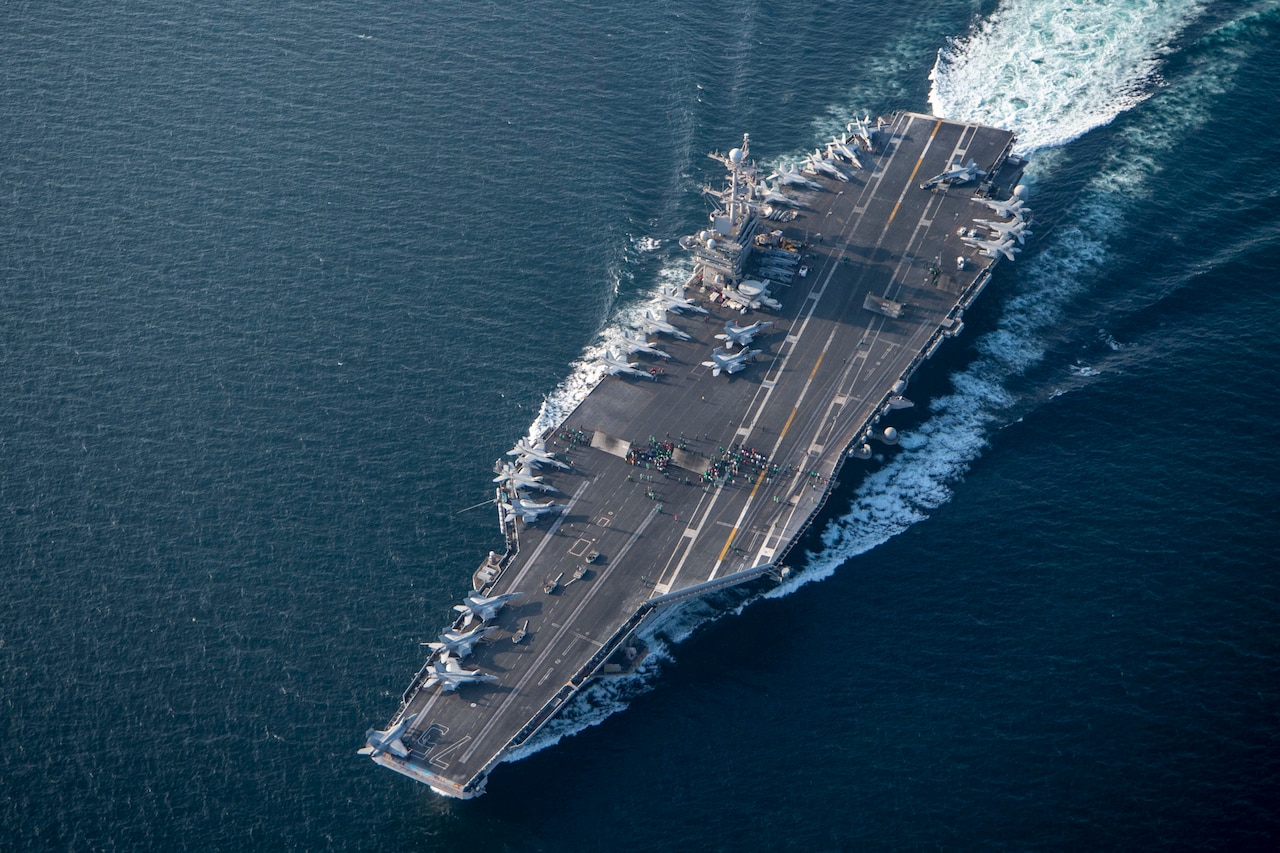 The flat surface of an aircraft carrier sailing on the ocean is seen in a photograph taken  from above.