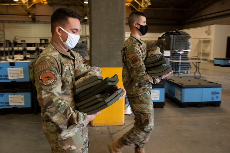 Two Airmen walking through a warehouse carrying heavy armor plates.