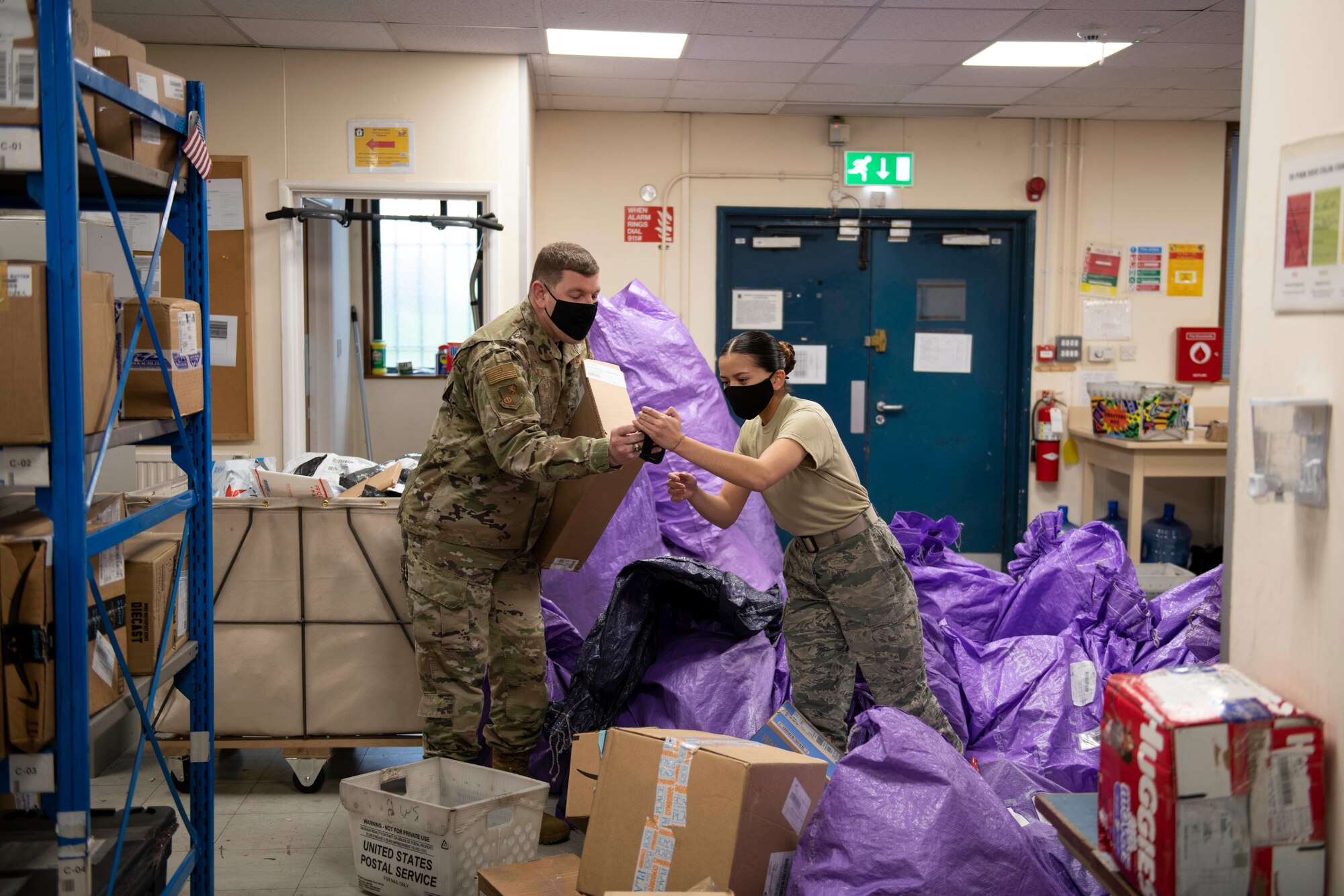 U.S. Air Force Airman 1st Class Melanie Macato, right, 423rd Force Support Squadron postal clerk, shows Col. Kurt Wendt, left, 501st Combat Support Wing commander, how to remove collected packages from the system, at the Royal Air Force Molesworth Post Office, England, Dec. 9, 2020. Airmen and civilians have volunteered at the post office to help with the influx of packages during the holiday season. (U.S. Air Force photo by Senior Airman Jennifer Zima)