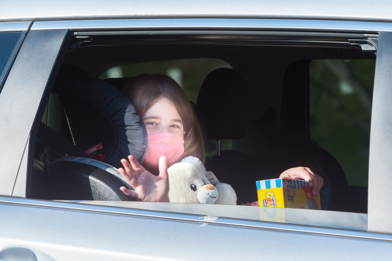A child in a car seat holds toys while waving out of her window.