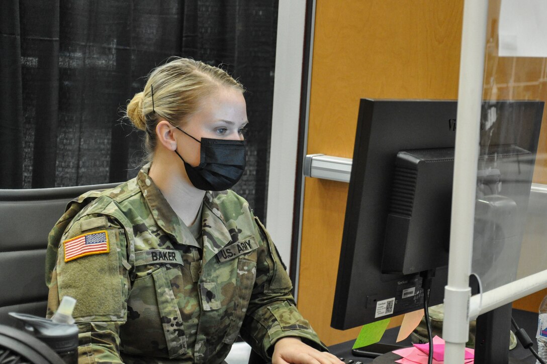 A soldier wearing a face mask sits at a desk looking at a computer