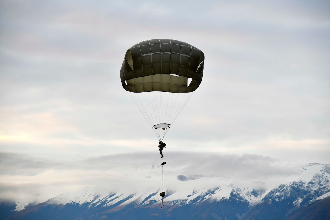 A soldier descends in the sky wearing a parachute with snowcapped mountains in the background.