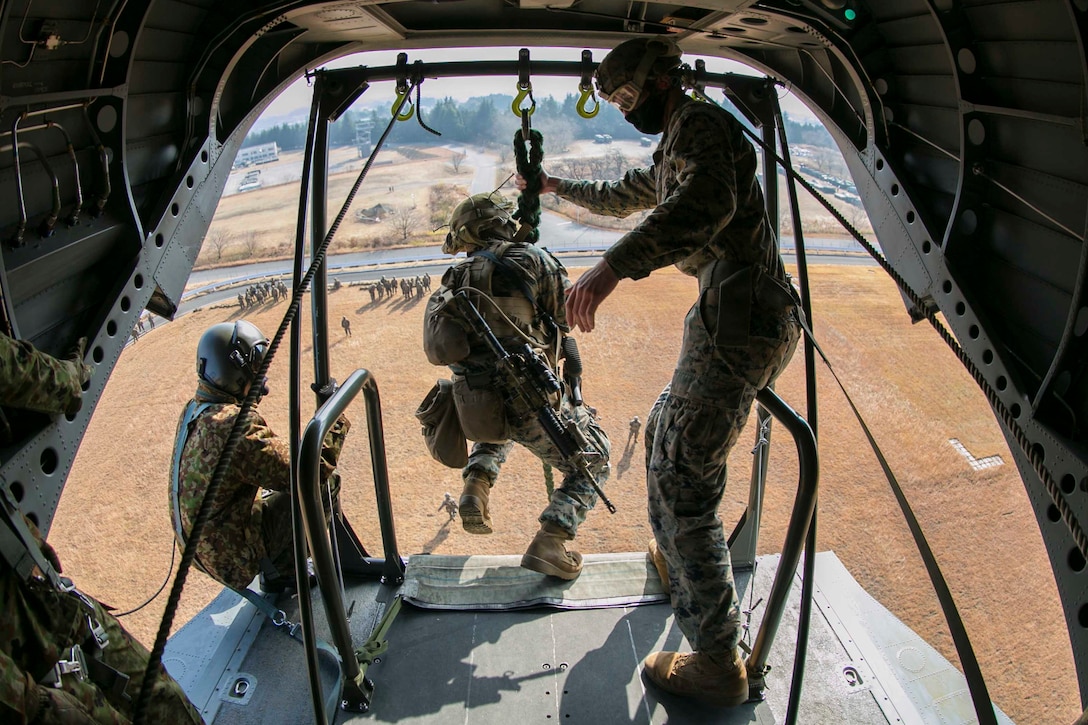 A Marine prepares to slide down a rope out of an aircraft as two others stand on either side.